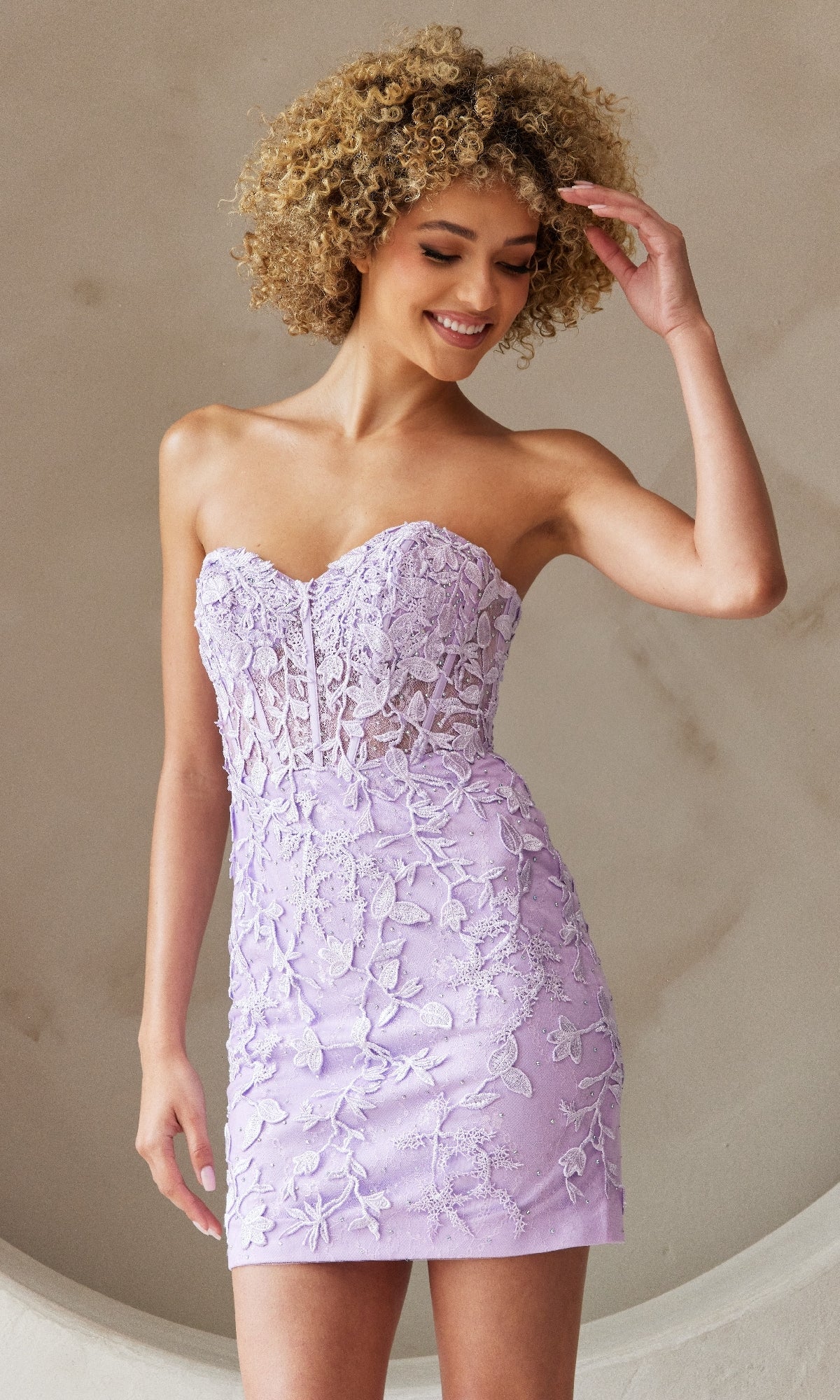 A woman in the Embroidered-Lace Strapless Short Hoco Dress 7053S with lilac floral lace stands against a neutral background, smiling and touching her hair.